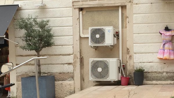 Deux climatiseurs split fixés sur la façade en pierre claire d’un immeuble, à côté d’un olivier en pot