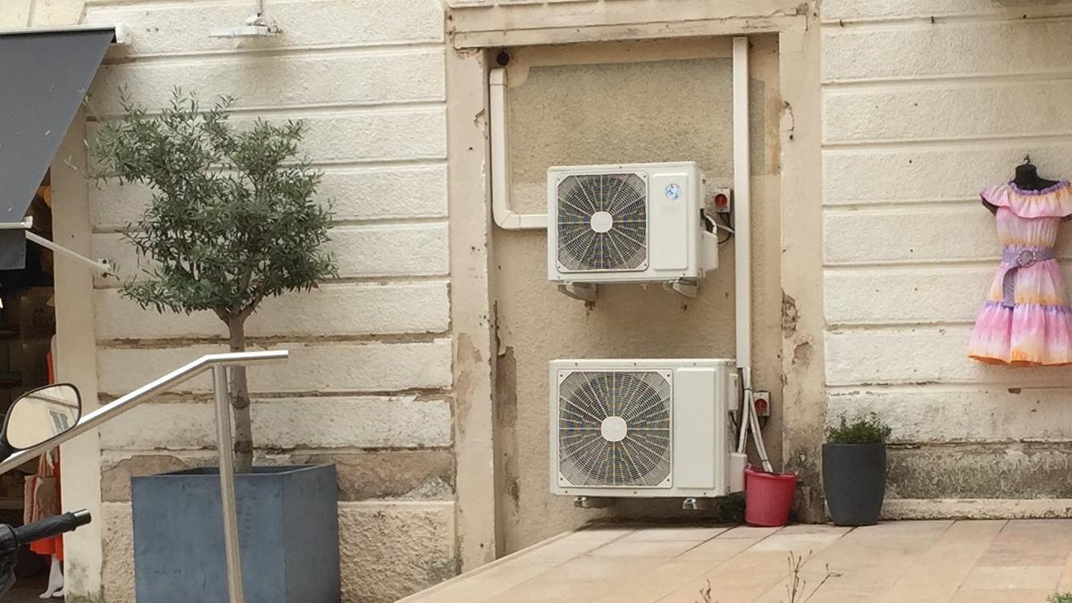 Deux climatiseurs split fixés sur la façade en pierre claire d’un immeuble, à côté d’un olivier en pot