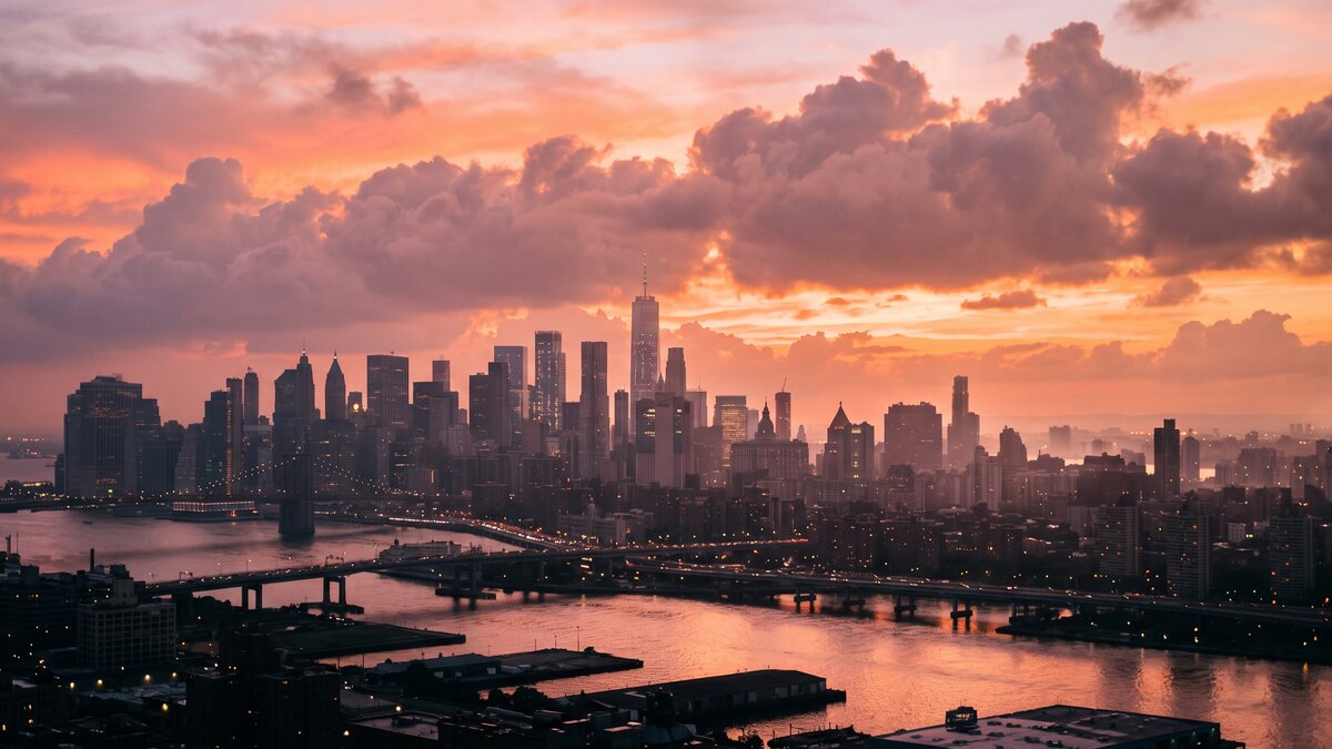 Silhouette d'un centre-ville dense au coucher du soleil, immeubles et une tour élancée se découpant sur un ciel orangé chargé de nuages, ambiance de chaleur persistante en fin de journée